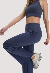 Frau in marineblauem Sportoutfit hebt ein Bein, trägt weiße und beige Sportschuhe, vor einem einfarbigen hellen Hintergrund.