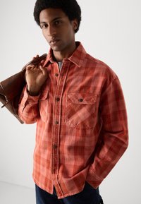 Camisa de franela de cuadros roja y naranja con dos bolsillos delanteros, cierre de botones y una textura suave. El modelo sostiene un bolso de cuero marrón.