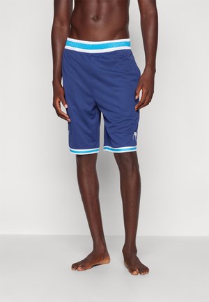 Man draagt blauwe sportshorts met een witte en turquoise tailleband en zoom, staat blootsvoets tegen een effen witte achtergrond.