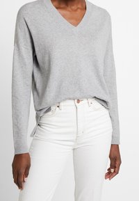 Šedý svetr s véčkovým výstřihem a dlouhými rukávy, měkké textury a bočními rozparky, v kombinaci s bílými džínami s vysokým pasem a zapínáním na knoflíky.