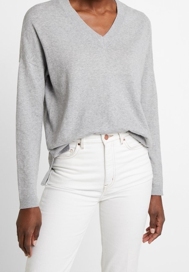 Šedý svetr s véčkovým výstřihem a dlouhými rukávy, měkké textury a bočními rozparky, v kombinaci s bílými džínami s vysokým pasem a zapínáním na knoflíky.