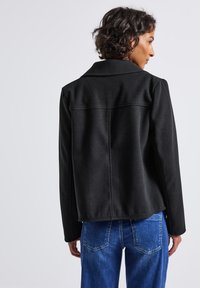 Black, structured jacket with a notched collar, textured fabric, and a slightly cropped design, paired with blue denim jeans.