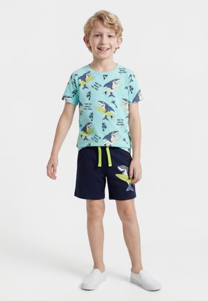 Dreng smilende og stående, iført en blå t-shirt med haj- og surfbrætmotiv samt marineblå shorts med hajgrafik og hvide slip-on sko.