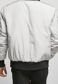 Man die een wit jack met lange mouwen draagt met zwarte manchetten en tailleband, gezien van achteren tegen een effen achtergrond.