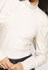 Cremfarbener Rollkragenpullover mit gerafftenÄrmeln, weich im Griff und figurbetontem Design, kombiniert mit dunklen Jeans. Verfügt über eine schlichte, minimalistische Silhouette.