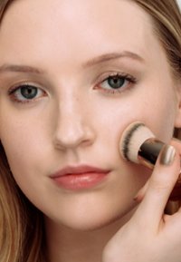 Foundationkwast die product op de wang aanbrengt. De kwast heeft een afgeronde vorm met zachte haren. Het model heeft een egale huid en lippen in een neutrale tint.