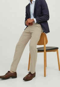 Beżowe dopasowane spodnie w zestawieniu z granatową marynarką i jasnoniebieską koszulą, uzupełnione brązowymi skórzanymi butami, siedzące na drewnianym krześle.