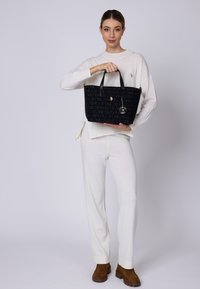 Sort tote bag med prægede logo mønster, gyldne detaljer og sorte håndtag, holdt af en person i en hvid outfit mod en lys baggrund.