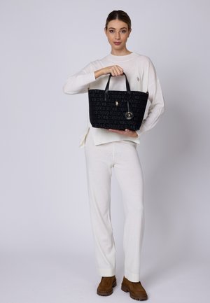 Svart tote-väska med präglat logomönster, guld-accenter och svarta handtag, hålls av en person i en vit outfit mot en ljus bakgrund.