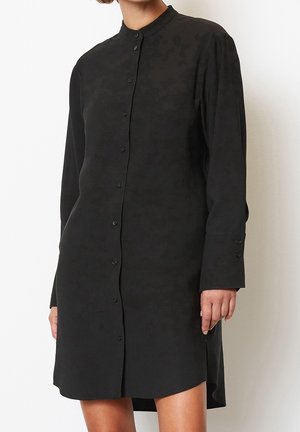 Czarna długa sukienka koszulowa z długim rękawem, zapięciem na guziki z przodu, kołnierzykiem mandaryńskim i lekko asymetrycznym dołem, noszona przez stojącą osobę.