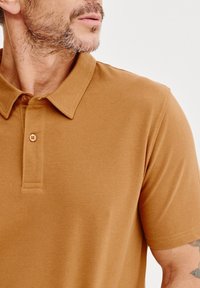 Koszulka polo z krótkim rękawem w ciepłym odcieniu brązu, wykonana z fakturowanej tkaniny, z kołnierzykiem, trzema guzikami i prostymi detalami ściegów.