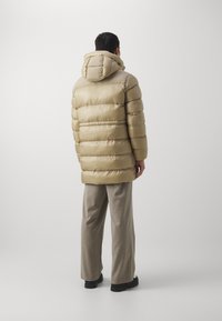 Beżowa kurtka puchowa z poziomym pikowaniem, kapturem i błyszczącym materiałem. W połączeniu z jasnobeżowymi spodniami i czarnym obuwiem.