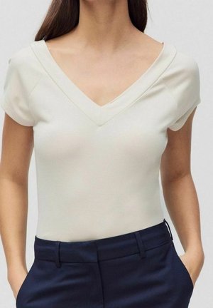 Woman wearing a short-sleeved, cream V-neck top tucked into high-waisted dark navy trousers with hands in pockets.