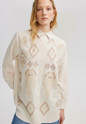 Camicia beige con bottoni caratterizzata da ricami geometrici in tonalità di marrone più chiaro, con colletto e maniche lunghe, realizzata in un tessuto morbido.