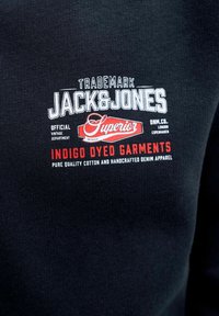 Tissu noir avec texte blanc et rouge indiquant "Jack & Jones Superior Indigo Dyed Garments pure quality cotton and handcrafted denim apparel."