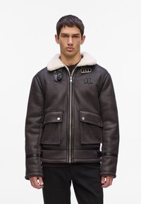 Chaqueta bomber de shearling marrón con un collar de piel sintética blanca, cierre de cremallera, dos bolsillos delanteros y un logotipo en el pecho. Acabado de cuero texturizado.