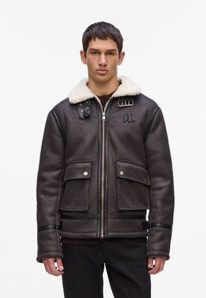 Brown shearling bomber jacket with a white faux fur collar, zip closure, two front pockets, and a logo on the chest. Textured leather finish.