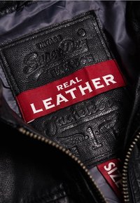 Interno di una giacca di pelle nera con un'etichetta rettangolare cucita. Etichetta rossa con la scritta "PELLE REALE" e dettagli del logo del marchio in rilievo.