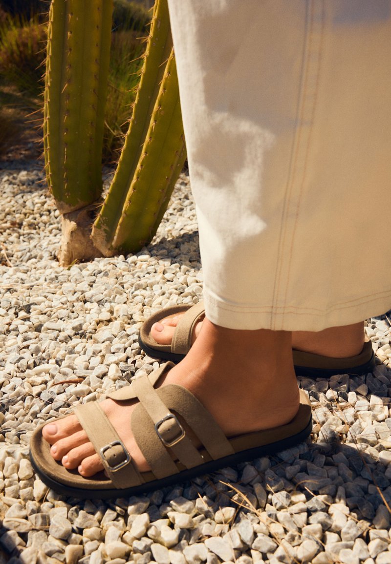 Personne portant des sandales en daim beige avec des brides à double boucle, debout sur du gravier blanc près d'un cactus vert.