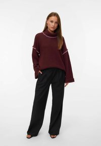Pull à col montant en tricot bourgogne avec des détails roses, associé à un pantalon noir à jambes larges. Coupe décontractée, tissu texturé, manches longues.