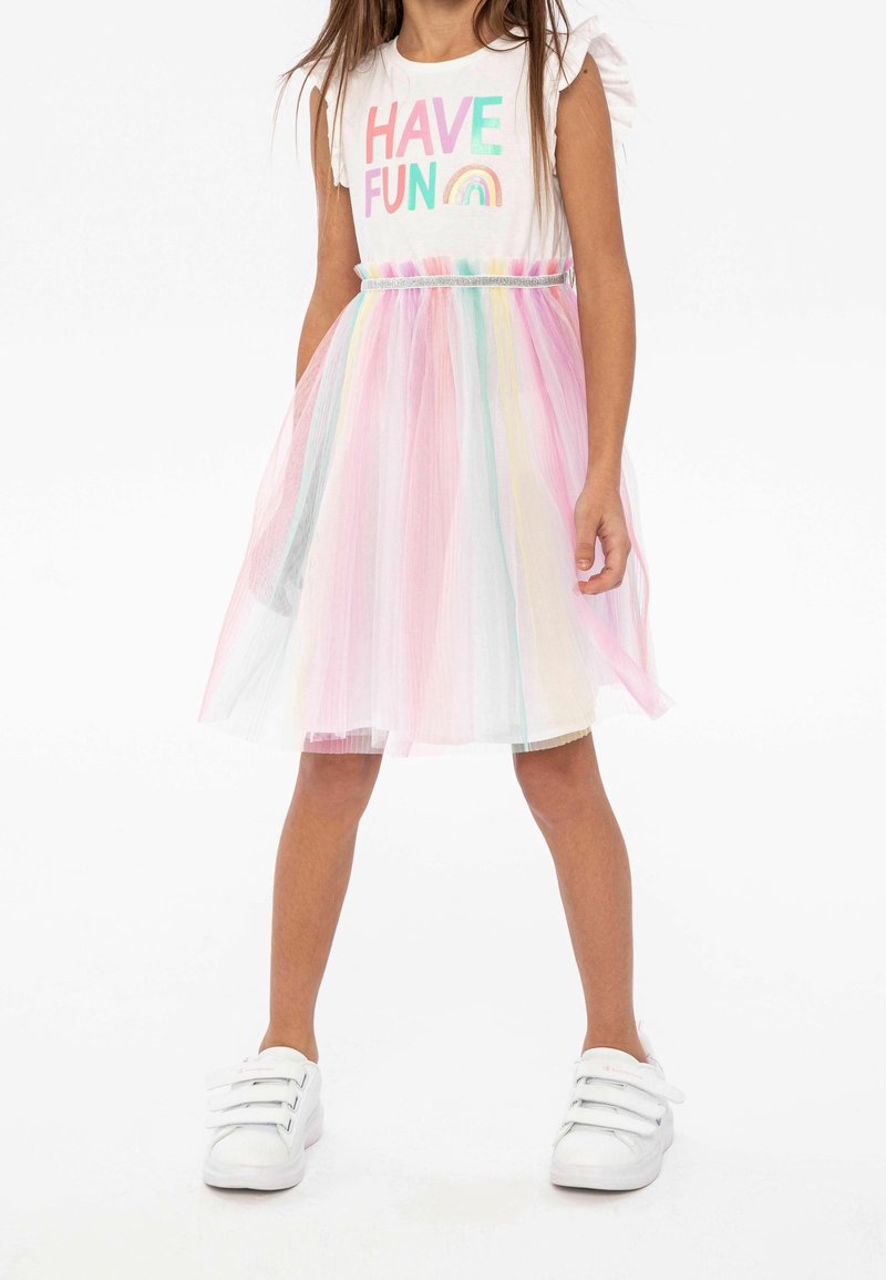 Fille portant une robe blanche avec le texte "Have Fun", une jupe en tulle arc-en-ciel pastel, et des baskets blanches à scratch, debout, jambes écartées.
