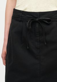 Κοντινό πλάνο μαύρης φούστας με δεμένη υφασμάτινη ζώνη στη μέση, φορεμένη με ανοιχτό μπεζ τοπ, που δείχνει μέρος του χεριού και του μπράτσου του φορέα.