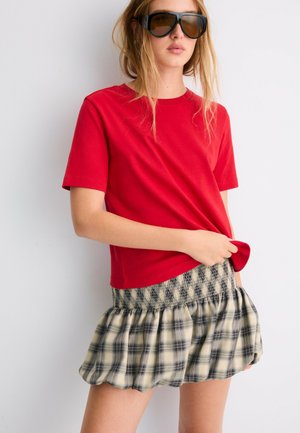 Ung kvinde iført oversize mørke solbriller, en rød kortærmet T-shirt og et kort, rynket ternet skørt står op ad en hvid væg.