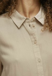 Beige, skjorte med krave lavet af letvægtsmateriale. Har en knaplukning foran med små, matchende knapper og en subtil tekstur.