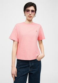 Νεαρό άτομο που φοράει ροζ μπλουζάκι με κοντά μανίκια και μικρό λογότυπο, σκούρο μπλε τζιν και ανοιχτό ροζ γυαλιά ηλίου, στέκεται με το ένα χέρι στην τσέπη.