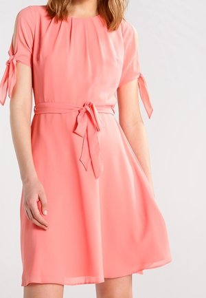 Žena nosící korálově růžové šaty ke kolenům s krátkými rukávy uvázanými na loktech a odpovídajícím páskem na mašli v pase.