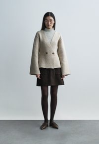 Beige wool blazer with wide sleeves and dual buttons, layered over a striped turtleneck. Dark brown suede skirt, black tights, and brown shoes.