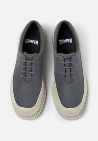 Zapatos grises con un acabado suave y suelas blancas contrastantes, con seis ojales y la marca "CAMPER" visible en la lengüeta y la plantilla.