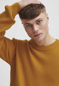 Textured mustard yellow sweater with ribbed pattern and crew neck. Model has short hair and smooth complexion, resting hand on head.