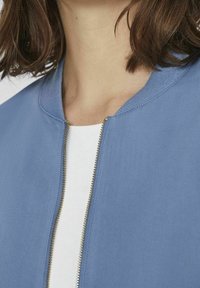 Light blue bomber jacket featuring a round neck, ribbed trim, and gold zipper, made from smooth fabric with a clean, simple design.