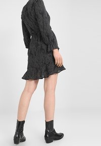 Mujer vistiendo un vestido negro de manga larga con estampado y dobladillo con volantes, con botas de cuero negras hasta la mitad de la pantorrilla, de pie frente a un fondo liso.