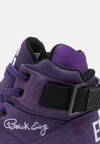 Fialové vysoké tenisky vyrobené z semišového materiálu, s texturovanými akcenty, výraznými tkaničkami a černým páskem s bílými logovými detaily.