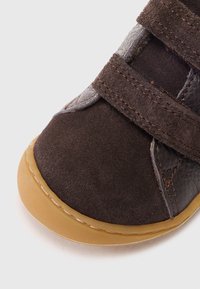 Shoe de camurça castanha com detalhes em couro, segurado por duas tiras de Velcro. Bico arredondado e sola de borracha flexível, apresentando uma borda exterior em tom bege.