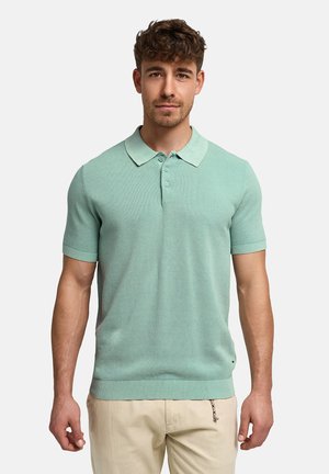 Homme aux cheveux courts et bouclés portant un polo à manches courtes vert menthe et un pantalon beige, debout devant un fond clair uni.