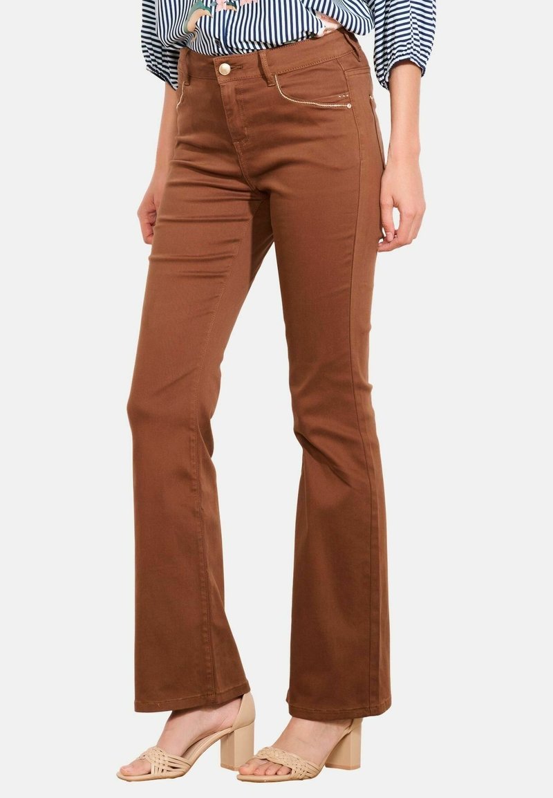 Femme portant un pantalon marron évasé, une chemise rayée et des sandales à talon beiges, debout devant un fond uni clair.