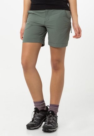 Kobieta w zielonych krótkich spodenkach turystycznych, fioletowych skarpetkach i czarnych butach trekkingowych, stojąca ze skrzyżowanymi nogami na białym tle.