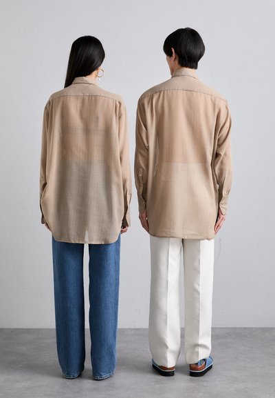 Camicie beige a bottoni con una consistenza morbida, dettagli di cucitura visibili e un taglio rilassato, abbinate a jeans blu e pantaloni bianchi.