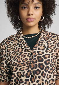 Camisa de manga corta con estampado de leopardo y una textura suave, con un logo negro, cierre de botones y cuello extendido.