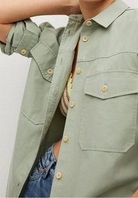 Chemise à manches courtes vert clair en tissu doux, dotée de deux poches poitrine, d'un denim déchiré et de boutons beiges. Les manches retroussées mettent en valeur son design.