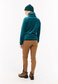 Mėlynas fliso megztinis su aukštu apykakle, derinamas su tan spalvos siaurais keliais, kuriuose yra užtrauktukai, ir tvirtais rudo spalvos žygio batais.