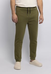 Olivgrüne Jogginghosen mit Kordelzugbund, Bündchen an den Knöcheln und seitlichen Taschen. Hergestellt aus weichem, strukturiertem Material. Kombiniert mit beigen Slippern.