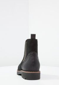 Bottine noire en cuir et daim, avec une forme incurvée, des perforations décoratives le long du bord et un talon bas et épais.