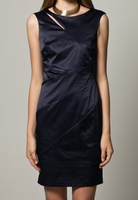 Mörkblå satin klänning med en åtsittande silhuett, med diagonala sömmar och ett distinkt axelutskärning, kombinerad med ett metalliskt halsband.