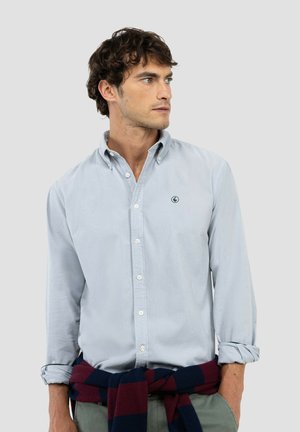 Hombre con cabello rizado que lleva una camisa azul claro abotonada y un suéter azul marino y burdeos atado alrededor de su cintura, mirando hacia la derecha.