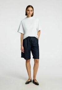 Valge lühikeste varrukatega T-särk kõrge kaelusega, koos tumedatest sinistest denim lühikestest pükstest, millel on mugav lõige ja volditud sääred. Mustad lamedad kingad.