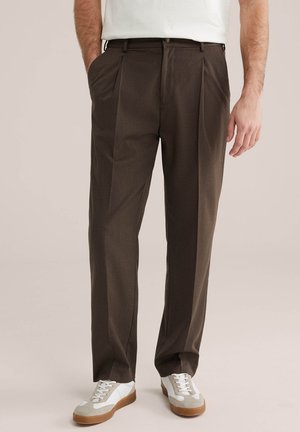 Muž oblečený v hnedých na mieru šitých nohaviciach s prednými záhybmi a bielych teniskách, stojaci s jednou rukou v vrecku.